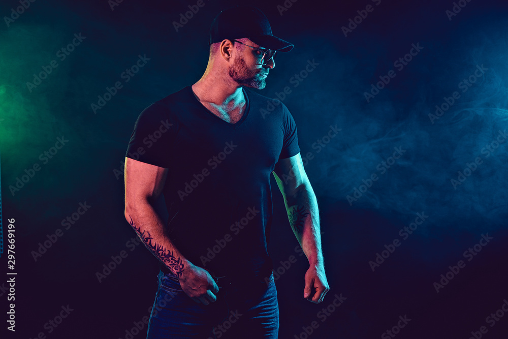 Fototapeta premium Young strong man bodybuilder in cap on wall background. Dark dramatic colors. Smoke on background.
