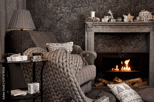 Modern cozy interior in gray shades with an armchair in front of fireplace. On the chair there are nice knitted blanket, cushions and Teddy bear. A lamp and gifts on the table. 
