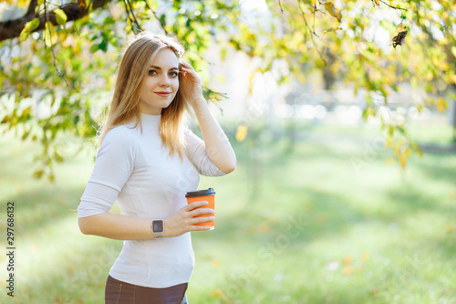 Young blonde woman enjoy autumn sunshine with a cup of coffee under tree at city park