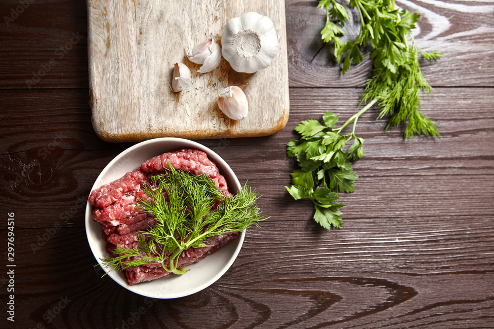 Bowl with raw minced meat, cutting board with fresh seasonings on a wooden background. White garlic heads and cloves, green dill and parsley leaves. Ground beef and condiments, top view Bowl with raw minced meat, cutting board with fresh seasonings on a wooden background. White garlic heads and cloves, green dill and parsley leaves. Ground beef and condiments, top view