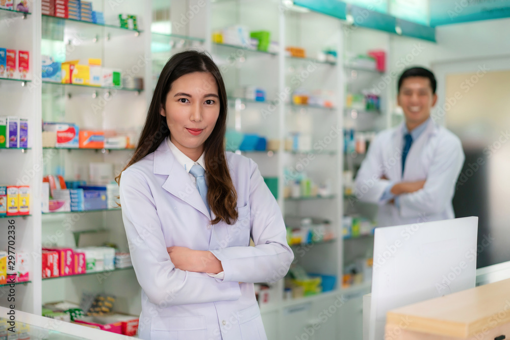 Two Asian young woman and man pharmacist with a lovely friendly smile standing with folded arms in the pharmacy drugstore. Medicine, pharmaceutics, health care and people concept.