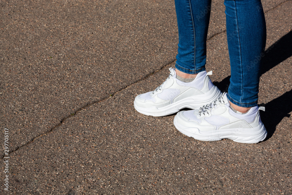 women's legs in white sports running shoes and jeans on the background of dry asphalt