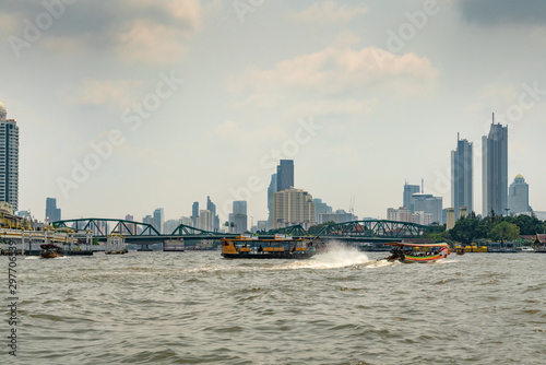Bangkok city, Thailand - March 17, 2019: Chao Phraya River. Wide view on metal Memorial Bridge over choppy water with small and larger ferry vessels. Skyline of skyscrapers under light blue cloudscape © Klodien