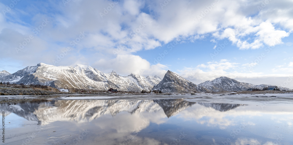 Panorama reflection of snowy mountains, blue sky and clouds on the calm clear water on a beach. A range of pointy mountain in beautiful winter landscape of Lofoten in northern Norway. Desolated land. 