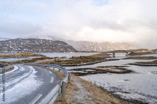 A long Atlantic Ocean winter Road and curved concrete bridge and water crossing from island in Lofoten. Sunshine on the far cloudy sky of bright future. Road barrier / guard rails for vehicle safety. 