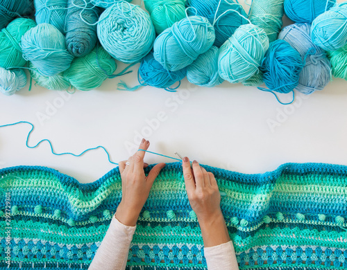 Women's hands are large. Woman crochets. Yarn of green, turquoise, aquamarine and blue colors. White wood background. photo