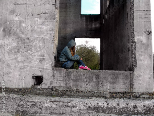 A teenage girl in a hood without a face sits in a rectangular hole of a concrete wall. The concept of teenage problems, a homeless child © ioanna_alexa