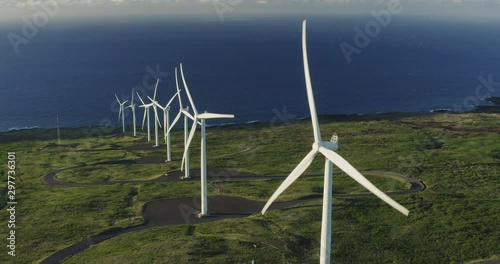 Aerial orbiting shot of a row of windmills spinning at sunrise with green fields and ocean in the background, wind energy farm generating electricity, green energy future photo