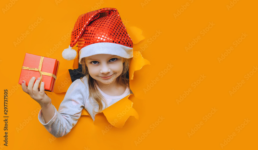 A beautiful little smiling girl in a red Santa hat looks out of a hole in orange paper and holds a box with a gift in her hand. Christmas and New Year concept. Bright coloured background, copy space A beautiful little smiling girl in a red Santa hat looks out of a hole in orange paper and holds a box with a gift in her hand. Christmas and New Year concept. Bright coloured background, copy space