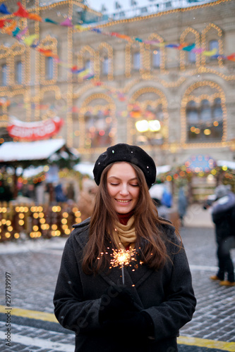 Attractive european lady in beret celebrating new year on the street, holding Bengal light. Outdoor portrait of happy brunette girl posing with sparkler in winter.