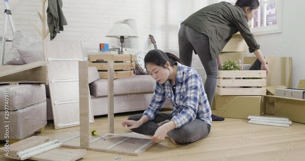 slow motion girl on floor reading instruction of diy wood product keep nodding head while taking screw understand assembly furniture. woman roommate in back move boxes unpacking relocation new house