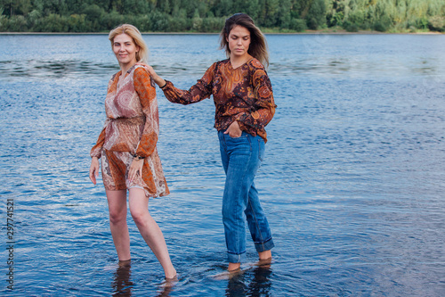 Two pretty sisters standing in river next to the shore on a cloudy windy summer day, having fun and laughing. photo