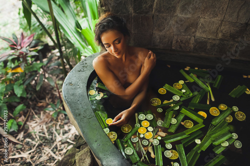 Beautiful young woman enjoying in outdoor spa. Luxury stone bath tub with jungle view. Natural organic tropical ingredients in the water: ginger, lime, orange and sea salt. Beauty treatment concept.
