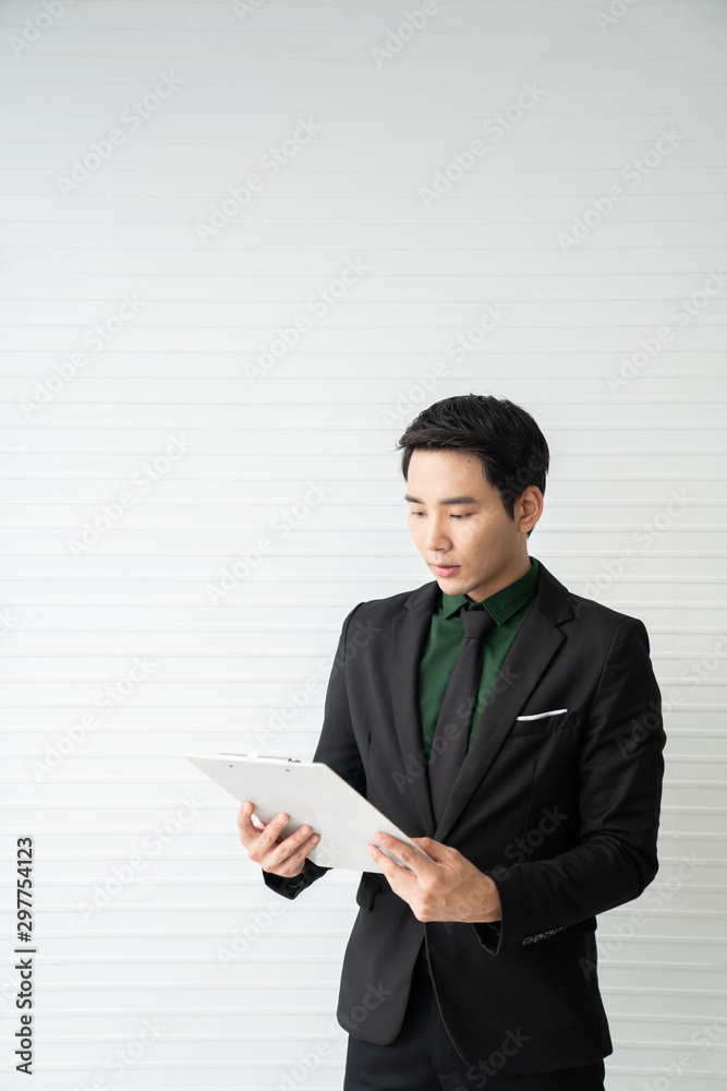 Asian businessman looking at report paper in his hand while standing inthe room with white wooden stripe background wall/ business concept / copy space Asian businessman looking at report paper in his hand while standing inthe room with white wooden stripe background wall/ business concept / copy space