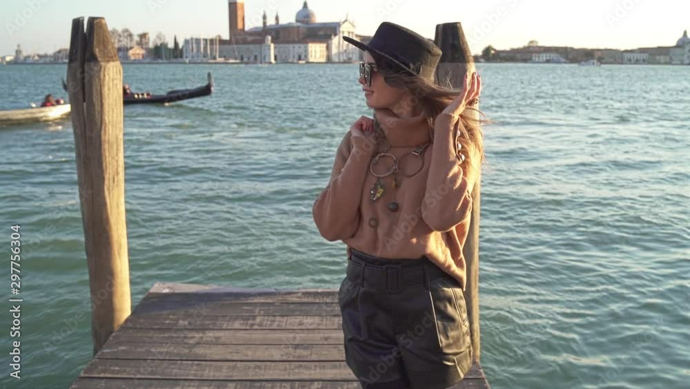 A remote shot of trendy smilled girl in black hat, glasses and brown sweater with brown hair who is watching into camera with blue sea, gondolas and old buildings on the background in Venice A remote shot of trendy smilled girl in black hat, glasses and brown sweater with brown hair who is watching into camera with blue sea, gondolas and old buildings on the background in Venice