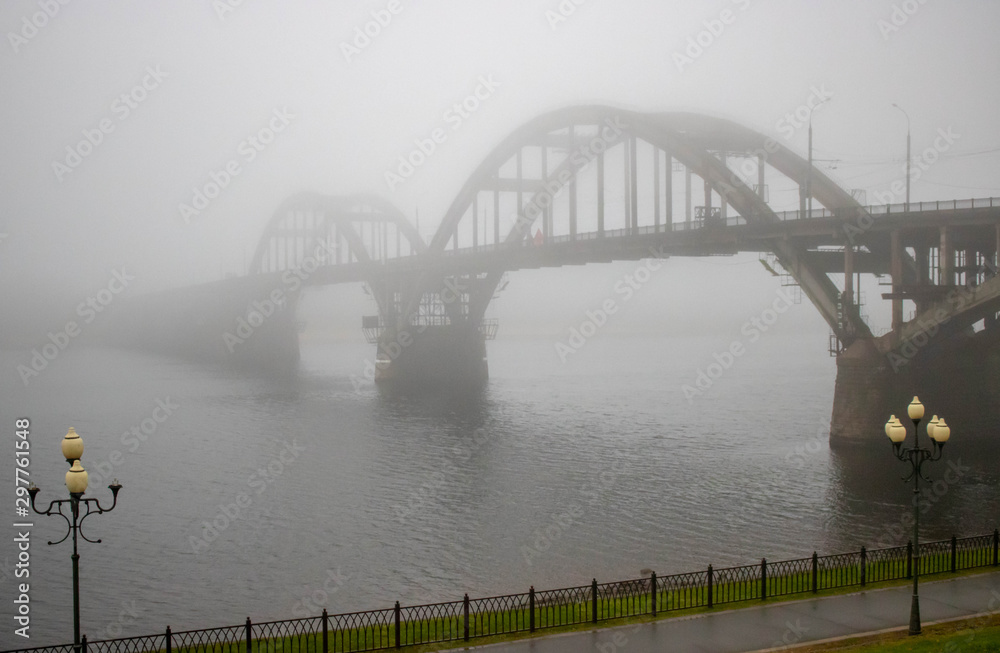 Obraz premium Autumn landscape with a riverbank strewn with fallen yellow leaves and a bridge in the fog.
