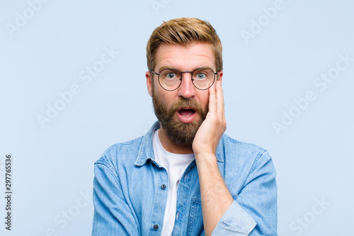 young blonde adult man open-mouthed in shock and disbelief, with hand on cheek and arm crossed, feeling stupefied and amazed photo