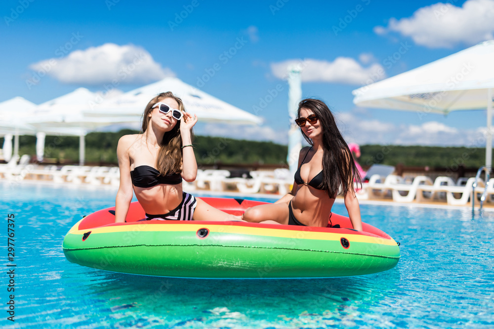 Attractive young girls in swimwear smiling while floating on the inflatable watermelon in the swimming pool.