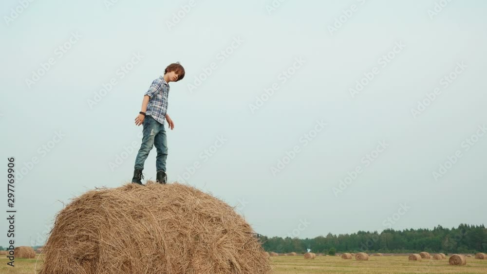 Stylish teenage boy posing on haystack in field. Low angle view of teenage boy in checkered shirt, jeans and leather boots standing, jumping and sitting on hay in countryside Stylish teenage boy posing on haystack in field. Low angle view of teenage boy in checkered shirt, jeans and leather boots standing, jumping and sitting on hay in countryside