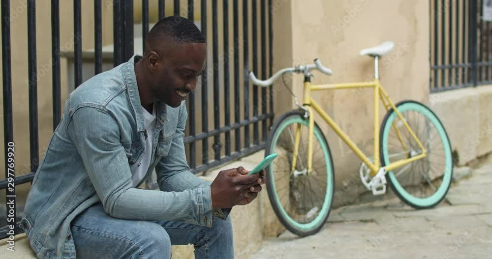 Happy African American Young Guy Using his Smartphone, Typing Messages, Chatting with Friends while Sitting near Stylish Bike with Old Metal Fance at the Background.