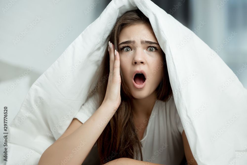 young beautiful brunette woman sitting in bed with blanket on her head looking amazed in the morning young beautiful brunette woman sitting in bed with blanket on her head looking amazed in the morning
