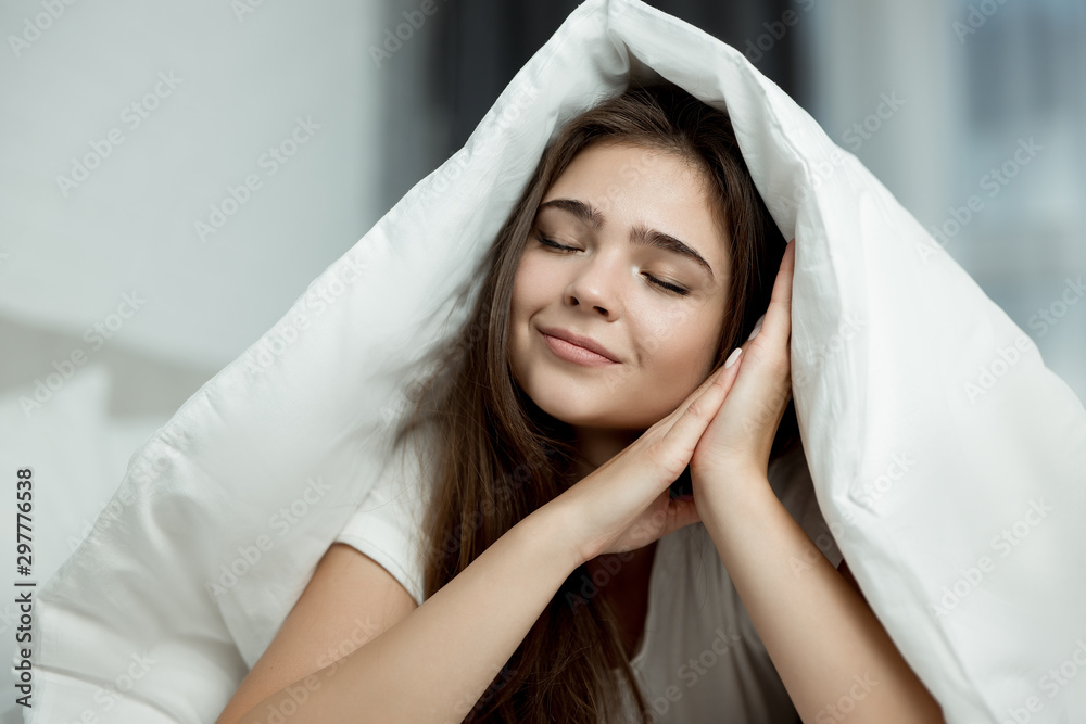 young beautiful brunette woman sitting in bed with blanket on her head with her hands near her face in the morning young beautiful brunette woman sitting in bed with blanket on her head with her hands near her face in the morning