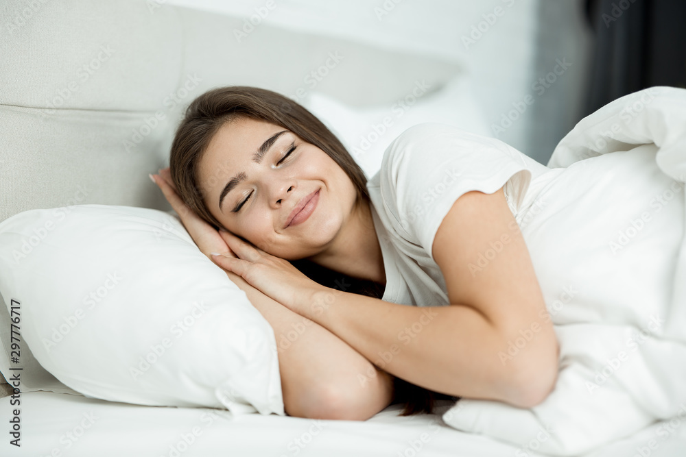 young attractive woman sleeping in bed in bright bedroom holding her hands near face in the morning young attractive woman sleeping in bed in bright bedroom holding her hands near face in the morning