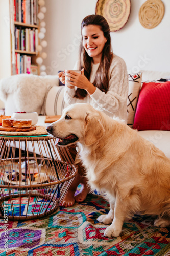 woman having a cup of tea at home during breakfast. Cute golden retriever dog besides. Healthy breakfast with fruits and sweets. lifestyle indoors © Eva