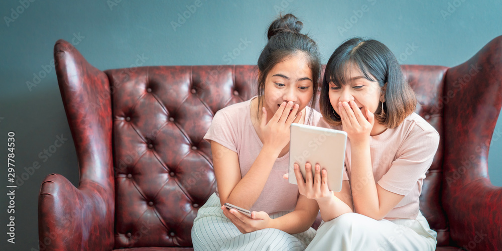 © sutthinon602 - Young asian women happy couple sitting on sofa shopping online using laptop a computer and smartphone in living room at home. Two young friends watching a video on a tablet in the living room.