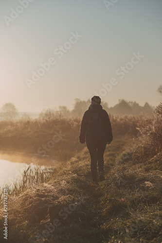 Woman walking through a meadow by a pond in the foggy morning. Sun rising above field and pond flooded with fog in the morning. Real people  authentic situations