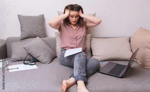 white woman sitting on a couch next to a laptop, document, woman with a scared, surprised expression, girl holding her head with her hands in fright