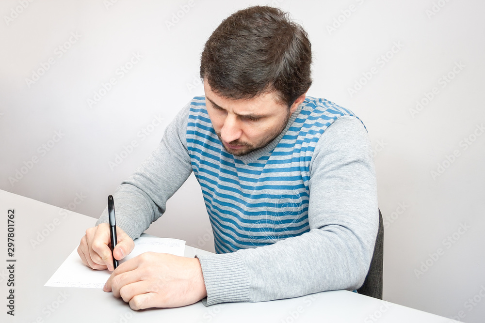 Handsome thoughtful bearded man in a striped sweater sits at the table and writes with a pen on paper