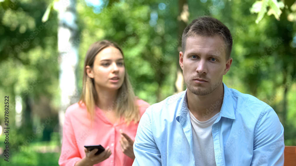 Sad young man looking camera, annoyed jealous girlfriend holding phone, distrust Sad young man looking camera, annoyed jealous girlfriend holding phone, distrust