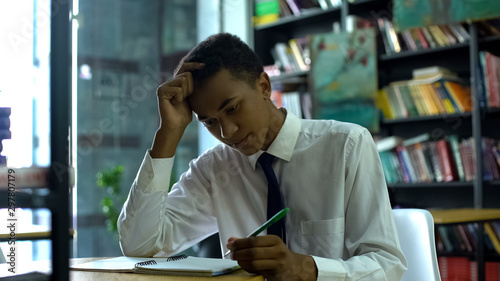 Black male high-schooler thinking about task, doing homework at campus library