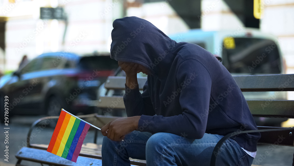 Black man in hood sitting on bench with lgbt minority flag, preconceptions Black man in hood sitting on bench with lgbt minority flag, preconceptions