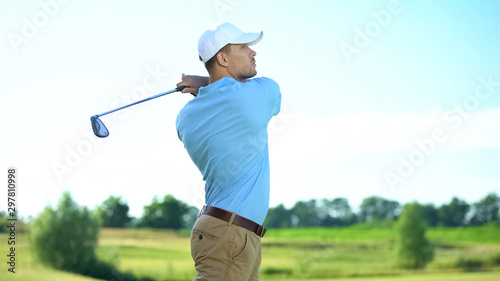 Professional golfer hitting ball in swing position, looking at meadow, sport