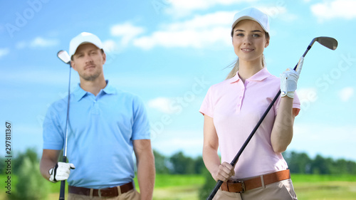 Young female golfer with partner holding clubs and smiling on camera, recreation