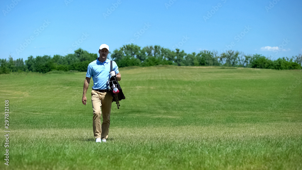 Experienced male coach moving on course with golf clubs bag, recreational hobby Experienced male coach moving on course with golf clubs bag, recreational hobby