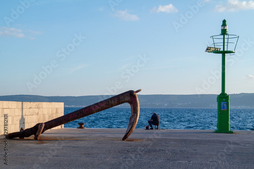 Lighthouse, big anchor and silhouette of the fisherman on the fisherman port.