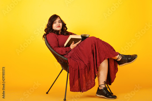 Attractive south asian woman in deep red gown dress posed at studio on yellow background sitting on chair with book. photo