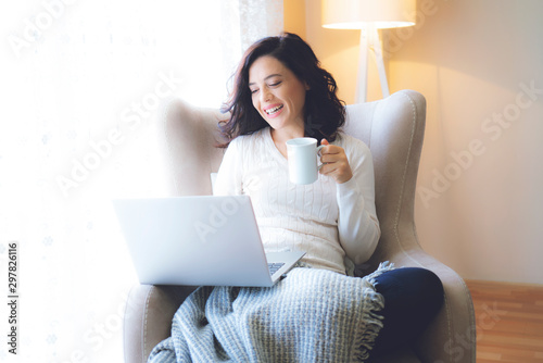 Enjoying time at home. Beautiful young smiling woman working on laptop and drinking coffee while sitting in a big comfortable chair at home photo