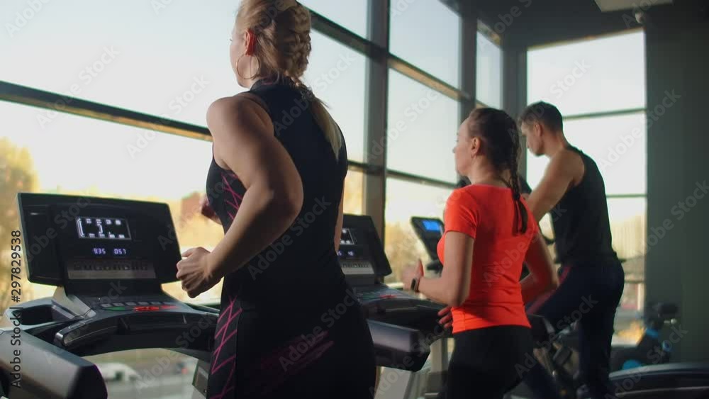A group of people running on a treadmill in a fitness room performing a cardio workout. Men and women train together Running indoors, warm-up before training in slow motion A group of people running on a treadmill in a fitness room performing a cardio workout. Men and women train together Running indoors, warm-up before training in slow motion