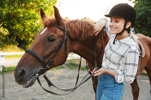 Image of blonde woman wearing hat standing by horse at yard in countryside