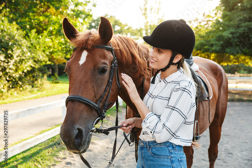 Image of pretty woman wearing hat standing by horse at yard in countryside