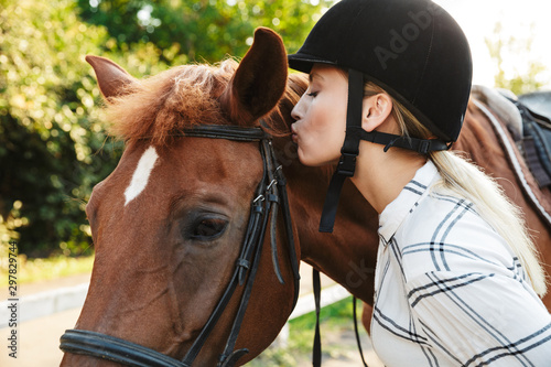 Image of tender woman wearing hat kissing horse at yard in countryside