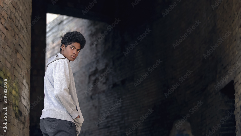 Black troubled teenager hanging around city building looking around, sadness