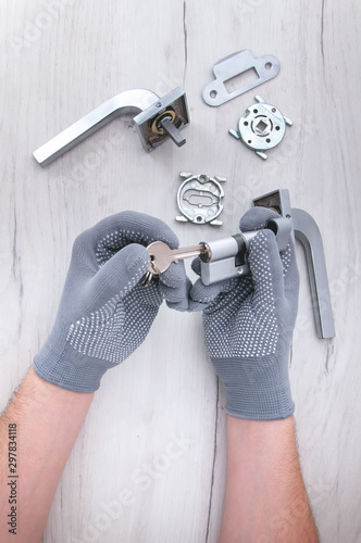 The guy holds in his hands in gray gloves the core of the lock and door keys on a light background. Repair in the house. Installing a new lock on the door. Spare parts for the door on a gray wooden ta