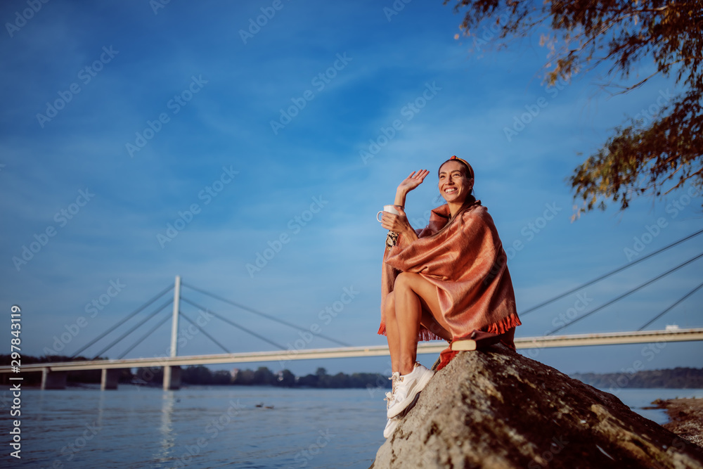 Charming brunette in floral dress and with headband sitting on rock on shore covered with blanket, waving to a friend and drinking beverage from mug. In background is bridge. Charming brunette in floral dress and with headband sitting on rock on shore covered with blanket, waving to a friend and drinking beverage from mug. In background is bridge.