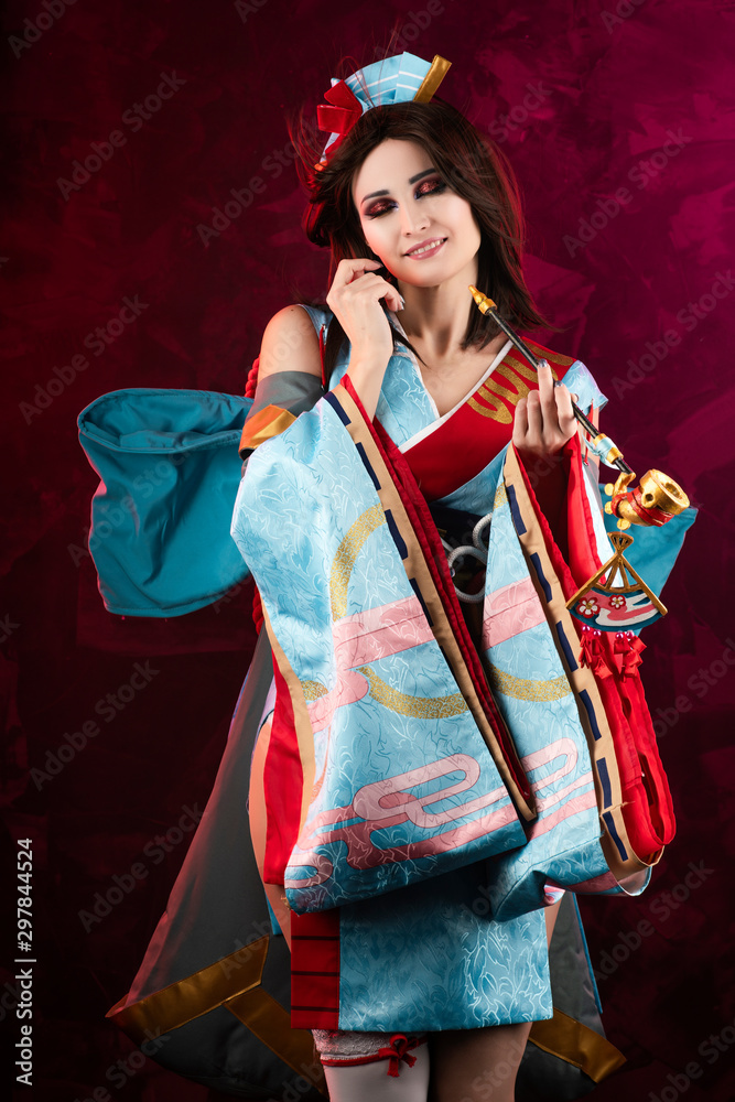 Beautiful leggy busty cosplayer girl wearing a stylized Japanese kimono  costume cheerfully posing holding a fake pipe on a blue background.  Stock-Foto | Adobe Stock