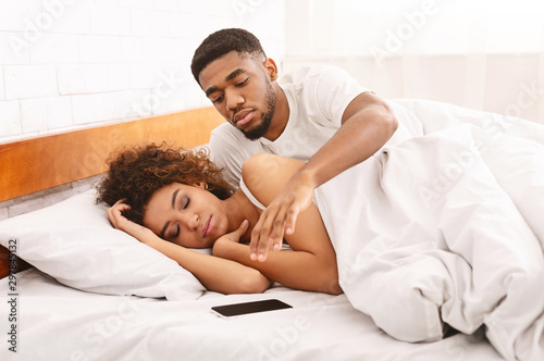 Fotografie Suspicious african man checking his sleeping wife's cellphone in bed